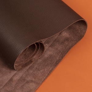 May include: A rolled sheet of dark brown leather, displaying a smooth, textured exterior. The underside reveals a softer, suede-like texture. The background is a solid orange colour, providing contrast to the leather.