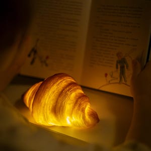 May include: A glowing, golden croissant illuminated from within, resting near an open book. The book's pages are visible, with illustrations and text. The scene is bathed in warm light, creating a cozy and inviting atmosphere.
