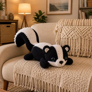 以下が含まれることがあります： 黒と白の毛糸で手編みされたスカンクのぬいぐるみ。大きな湾曲した尻尾、小さな丸い耳、暗いプラスチック製の安全な目を持つ。頭から背中にかけて白いストライプが入っており、横たわった姿勢で配置されている。