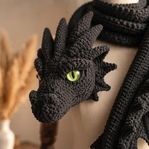 May include: A dark charcoal gray crochet scarf featuring a three-dimensional dragon head with prominent spikes and a textured, scaly appearance. The dragon head has a single visible bright green eye with a vertical black pupil, crafted in yarn.