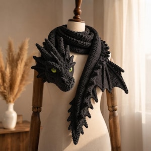 May include: A dark charcoal gray crocheted scarf shaped like a dragon. The scarf features a detailed dragon head with bright green eyes, a long body with a serrated spine, and a large, textured wing extending from the side of the neck area.