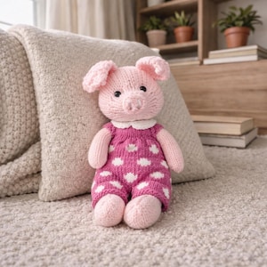 May include: A handmade pink knitted pig doll with a white collar and a pink jumpsuit patterned with white clouds. The pig has black eyes and pink ears, arms, and legs. The doll is sitting on a beige carpet in front of a beige pillow.