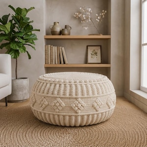 Può includere: Un pouf rotondo all'uncinetto color crema caratterizzato da un motivo geometrico testurizzato con forme a rombo in rilievo e dettagli a coste verticali lungo i lati. Il pezzo è realizzato in filato spesso e poggia a terra, offrendo una struttura cilindrica solida per sedersi o appoggiare i piedi in una stanza dai toni neutri.