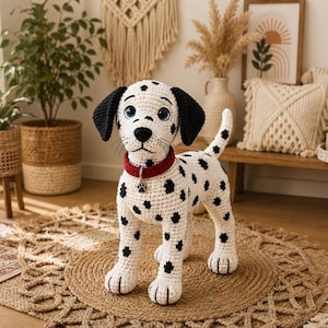 May include: A crocheted Dalmatian dog toy with white yarn and black spots, featuring large blue eyes, black ears, and a red collar with a small silver paw print charm. The stuffed animal stands on four legs with a raised tail and stitched details.