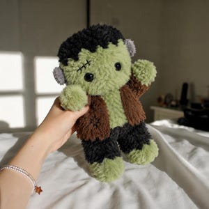 May include: A plush toy resembling a monster with fuzzy light green skin, black hair, and a brown jacket. The toy features black button eyes, small gray ears, and stitched details on the forehead and jacket, crafted from soft, textured fabric.