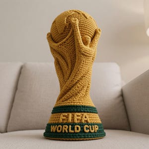 Op de afbeelding: Een goudgele gehaakte replica van de FIFA World Cup-trofee met een gestileerde menselijke figuur die een wereldbol omhoog houdt. De basis is donkergroen met de tekst FIFA WORLD CUP in goudgele letters op de voorzijde van de trofee.