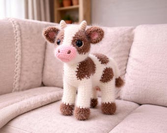 May include: A handmade crochet stuffed animal shaped like a cow. The cow has a white body with brown patches, a pink snout, small horns, and dark eyes. The yarn texture is fuzzy and soft, featuring four legs and a small tail.