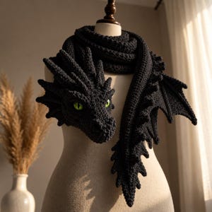 May include: A dark charcoal gray crocheted scarf shaped like a dragon, featuring a detailed head with bright green eyes, textured scales along the neck, and a long tail section with small wing-like appendages extending from the side of the scarf.