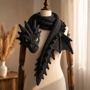 May include: A black crocheted scarf shaped like a dragon, featuring a detailed dragon head with bright green eyes, a long body with a serrated spine, and a large wing extending from the side, all displayed on a neutral-colored mannequin torso.