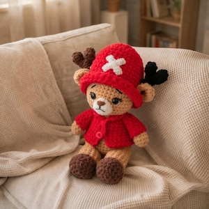 May include: A handmade crochet plush toy of a reindeer character, wearing a red hat with a white cross and a matching red jacket. The toy has brown antlers, black heart-shaped details, and brown feet. It is sitting on a beige couch.