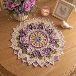 Op de afbeelding: Een rond, crèmekleurig gehaakt kleedje met een centraal wielpatroon omringd door een ring van paarse bloemen met witte parelcentra en groene bladeren, afgewerkt met een delicate, puntige, geschulpte rand in lichtpaars en crème.
