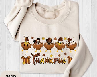 May include: Sand-colored sweatshirt featuring a Thanksgiving-themed design. The graphic includes cartoon characters dressed as turkeys, with the word "THANKFUL" in brown script. Autumn leaves and a pumpkin accent the design.