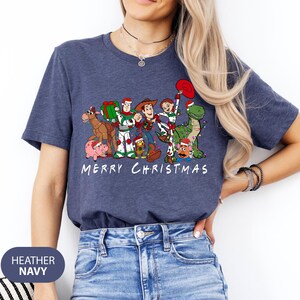 May include: Heather navy blue t-shirt featuring a graphic of Toy Story characters in Christmas attire. The design includes Woody, Buzz Lightyear, and others, with the text "MERRY CHRISTMAS" in white.