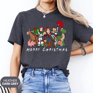 May include: Heather dark grey t-shirt featuring a graphic of Toy Story characters in Christmas attire. The design includes Woody, Buzz Lightyear, and other characters with the text "MERRY CHRISTMAS" in white.