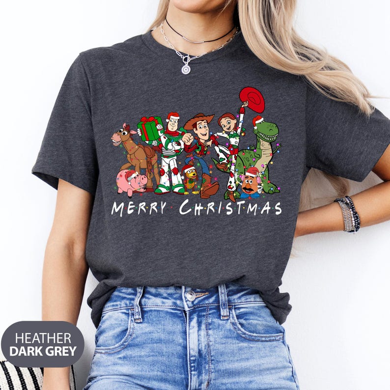 May include: Heather dark grey t-shirt featuring a graphic of Toy Story characters in Christmas attire. The design includes Woody, Buzz Lightyear, and other characters with the text "MERRY CHRISTMAS" in white.