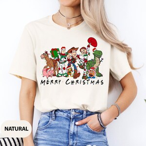 May include: Cream-colored t-shirt featuring a graphic of animated characters, each adorned with Christmas lights and accessories. The text "MERRY CHRISTMAS" is printed below the characters. The shirt is made of a soft material.