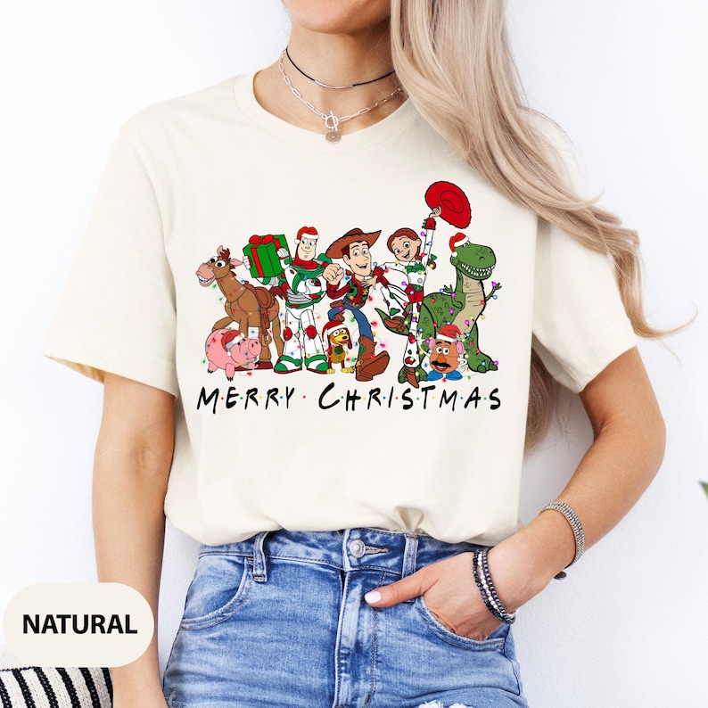 May include: Cream-colored t-shirt featuring a graphic of animated characters, each adorned with Christmas lights and accessories. The text "MERRY CHRISTMAS" is printed below the characters. The shirt is made of a soft material.
