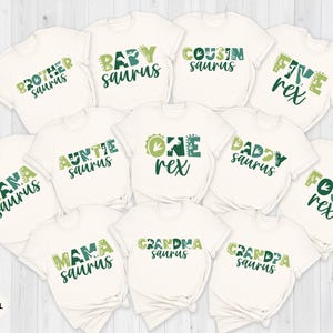 May include: A collection of off-white t-shirts with various green text designs. The shirts feature phrases like "Brother Saurus," "Baby Saurus," and "One Rex." The text has a dinosaur theme.