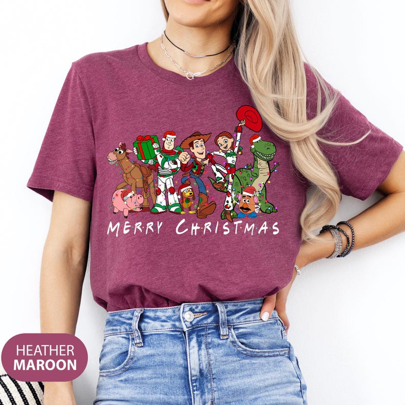 May include: Heather maroon t-shirt featuring a graphic of Toy Story characters with Christmas decorations. The text "MERRY CHRISTMAS" is printed in white below the characters. The shirt is a crew neck and has short sleeves.