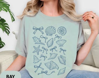 May include: A light blue t-shirt featuring a collection of blue seashell illustrations and bows. The word "BAY" is printed on a light green oval at the bottom left of the shirt. The shirt is made of a soft material.