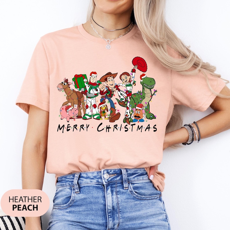 May include: Heather peach-colored t-shirt featuring a graphic of Toy Story characters with Christmas decorations. The text "MERRY CHRISTMAS" is printed below the characters. The shirt is a short-sleeved crew neck.