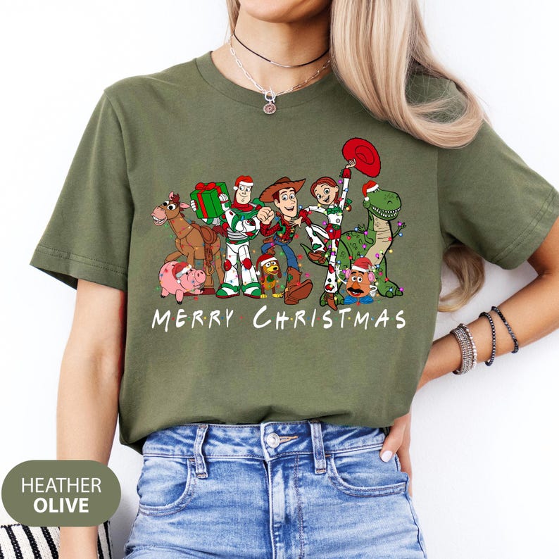 May include: Heather olive green t-shirt featuring a graphic of animated characters in Christmas attire. The characters include a cowboy, a dinosaur, and other figures. The text "MERRY CHRISTMAS" is printed below the graphic.