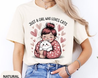 May include: Cream-colored t-shirt featuring a graphic of a girl holding a white cat, surrounded by pink hearts. The text "JUST A GIRL WHO LOVES CATS" is printed above the illustration. The shirt is made of a soft material.