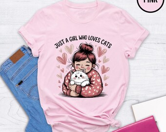 May include: Pink t-shirt with the text "JUST A GIRL WHO LOVES CATS" and a graphic of a girl holding a white cat, surrounded by hearts. The shirt is displayed on a light wood surface with blue jeans and a pink wallet.