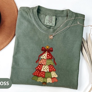 May include: A moss green t-shirt with a Christmas tree design. The tree is made of patchwork squares in red, green, and white patterns, topped with a red bow and gold star. A silver necklace with a small pendant is also visible.