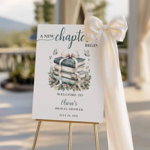May include: A welcome sign for a bridal shower, featuring a stack of books with floral accents and butterflies. The sign reads "A New Chapter Begin" and "Welcome to Olivia's Bridal Shower, July 24, 2025." The sign is displayed on a gold easel and adorned with a cream-colored bow.