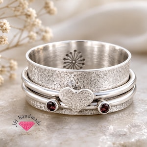 May include: A silver spinner ring featuring a wide, textured band with a central heart-shaped charm and two small, round, dark red garnet stones. The interior of the band displays a stamped starburst pattern. The text JH handmade jewellery is visible.