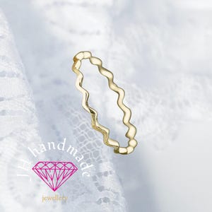 Puede incluir: Un delicado anillo dorado con un diseño ondulado. El anillo está sobre un fondo de encaje blanco. El diseño del anillo es un patrón continuo y ondulado. La imagen también incluye el texto "JH handmade jewellery" con un logotipo de diamante rosa.