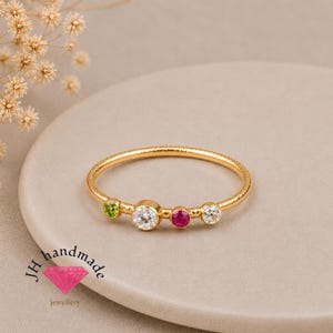 May include: A gold ring with a textured band featuring four bezel-set stones: a small green peridot, a large clear white diamond-like stone, a medium pink ruby-like stone, and a small clear white stone, with the text JH handmade below.