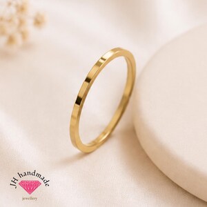 May include: A thin, polished gold band ring with a smooth, flat surface. The ring is positioned at an angle, showcasing its circular shape and metallic finish. The text JH handmade jewellery with a pink diamond icon is visible in the corner.