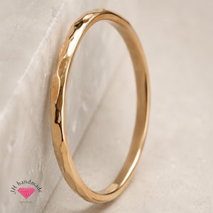 Könnte beinhalten: Ein dünner, kreisförmiger Goldring mit einer gehämmerten, texturierten Oberfläche. Das Band hat eine reflektierende, polierte Oberfläche, die das Licht einfängt. Das Logo JH handmade jewellery mit einem kleinen rosa Diamantsymbol ist unten links zu sehen.