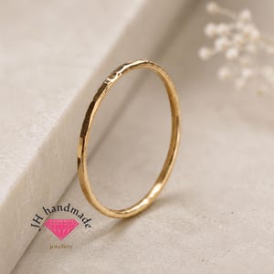 May include: A thin, gold-coloured metal ring with a hammered, textured surface. The ring features a circular band design. The text JH handmade is printed in black with a pink diamond-shaped icon located below the text on the lower left side.