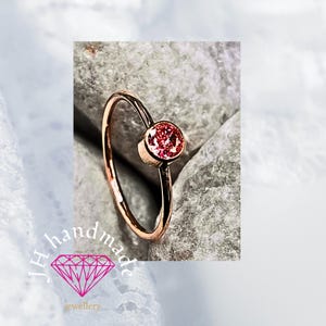 Op de afbeelding: Een roségouden ring met een ronde, roze edelsteen. De ring is geplaatst op een grijze stenen ondergrond. De afbeelding bevat de tekst "JH handmade jewellery" met een diamantafbeelding.