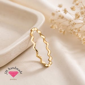 Può includere: Un anello in oro caratterizzato da un design a fascia continua ondulata e serpentina. L'anello presenta una finitura metallica liscia e lucida. Il logo di JH handmade jewellery, con un'icona di diamante rosa e testo, è visibile in basso.