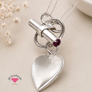 May include: A silver-toned necklace featuring a toggle clasp with a cylindrical bar and a circular ring. A single deep red gemstone is set into the end of the bar. A brushed silver heart-shaped pendant hangs from the toggle. Text reads JH handmade.