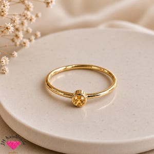 Op de afbeelding: Een dunne gouden ring met een enkele, ronde, geelkleurige edelsteen in een gladde zetting. De ringband is glad en gepolijst en heeft een eenvoudig ontwerp met een centrale steen die het licht reflecteert. De ring ligt op een lichtgekleurd oppervlak.