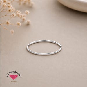 Puede incluir: Un anillo de banda de plata pulida y fina con una superficie lisa y redondeada. El anillo es circular y presenta un acabado metálico reflectante. En la esquina inferior izquierda, hay un logotipo con el texto JH handmade y un pequeño icono de diamante rosa.