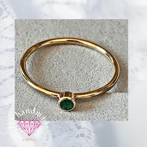 May include: A slender gold ring featuring a small, circular, emerald-green gemstone. The ring has a thin band and a simple, elegant design. The ring is displayed on a light grey surface.