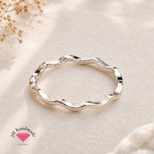 Puede incluir: Un anillo de plata pulida y fina que presenta un diseño de banda continua ondulada o con ondas. La superficie del metal es lisa y reflectante, captando la luz a lo largo de sus bordes ondulados. El anillo se presenta como una banda circular.