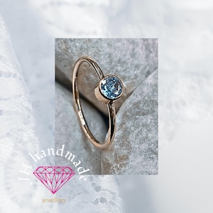 May include: A gold ring with a light blue gemstone, set in a bezel. The ring has a slender band. The gemstone is round. The ring is displayed on a light grey stone surface. The image includes the text "JH handmade jewellery".