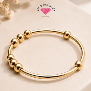 Könnte beinhalten: Ein goldfarbener Armreif mit glatter, polierter Oberfläche und sechs gleichmäßig verteilten, kugelförmigen Goldperlen entlang des kreisförmigen Bandes. Das Logo JH handmade jewellery mit einem rosa Diamantsymbol ist über dem Armreif zu sehen.