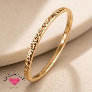 May include: A thin gold ring with a textured, hammered surface finish. The band features a consistent, rugged pattern along its outer edge. The logo for JH handmade jewellery, featuring a pink diamond icon, is visible in the bottom left corner.