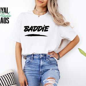 May include: White t-shirt with the word "BADDIE" in black, underlined with a black brushstroke. The shirt also features the text "ROYAL Studio CHAOS" in black and green. The model is wearing blue jeans.