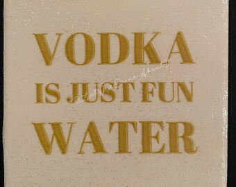 May include: A white square coaster with gold glitter and the text "VODKA IS JUST FUN WATER" in gold lettering.