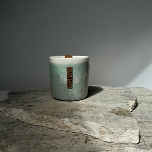 Könnte beinhalten: Eine zylindrische Kerze mit weißem Oberteil und hellblauem, strukturiertem Körper. Ein rechteckiges Holzschild mit dem Wort "malmo" ist an der Kerze befestigt. Die Kerze steht auf einer grauen, strukturierten Steinoberfläche.