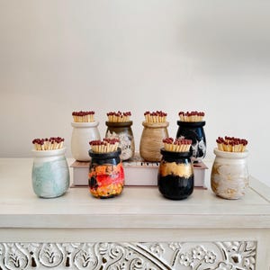 May include: A selection of decorative match holders in various colours and patterns. Each small ceramic jar is filled with red-tipped matches. The jars are displayed on a white table with carved details, creating a visually appealing arrangement.